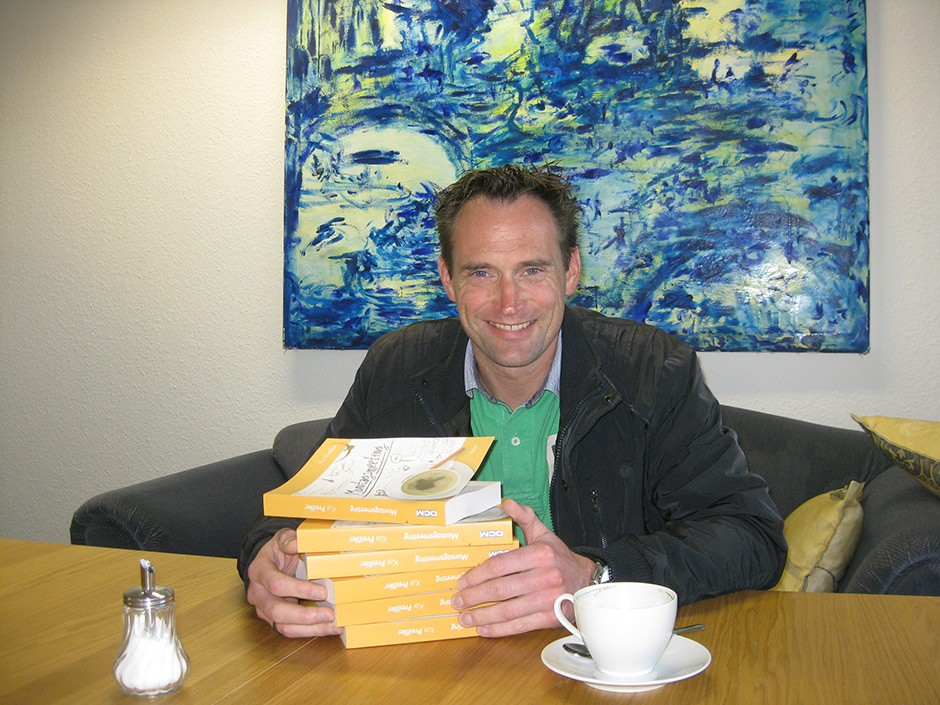 Kai Preißler sitzt auf dem blauen Sofa im Verlag. Vor ihm ein großer Bücherstapel, den er mit den Händen umfasst.
