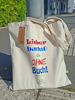 Stehende beige Bio-Baumwolltasche im Freien auf grauem Asphalt vor grünen Hecken. Der bunte Spruch "Lieber barfuß als ohne Buch!" ist gut sichtbar auf der Vorderseite aufgedruckt.